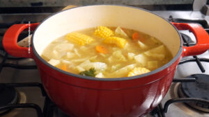 A red pot sits on a stove, filled with a simmering Mexican vegetable soup featuring corn on the cob pieces, chopped carrots, potatoes, cabbage, and other vegetables in a clear broth.
