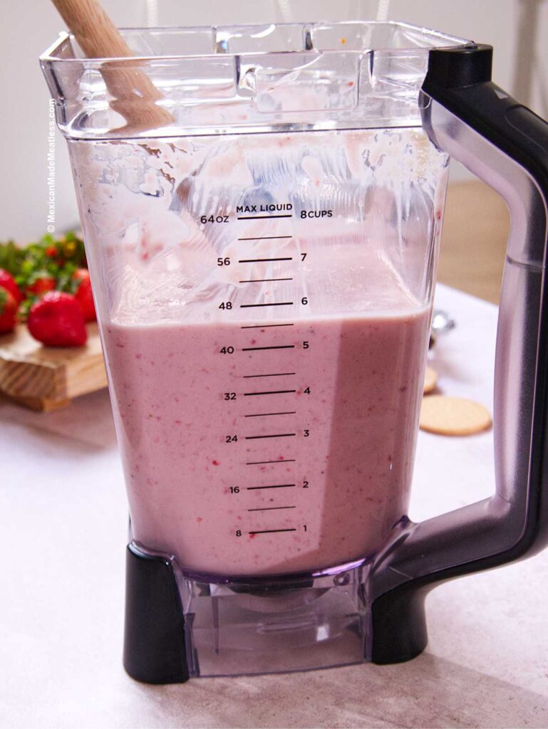 A blender pitcher filled with pink strawberry cream to make Carlota de Fresa, marked with measurements up to 64 ounces. A wooden spoon rests inside, and fresh strawberries are visible on a cutting board in the background.