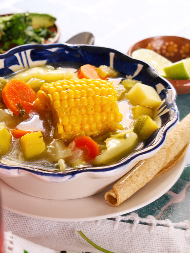 A bowl of Mexican vegetable soup with corn on the cob, carrots, potatoes, and zucchini, served with rolled tortillas. Lime slices and chopped cilantro are in the background on a white tablecloth.
