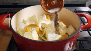 A hand pours salt and pepper from a small bowl into a red pot filled with chopped cabbage and corn on the cob, placed on a stovetop.