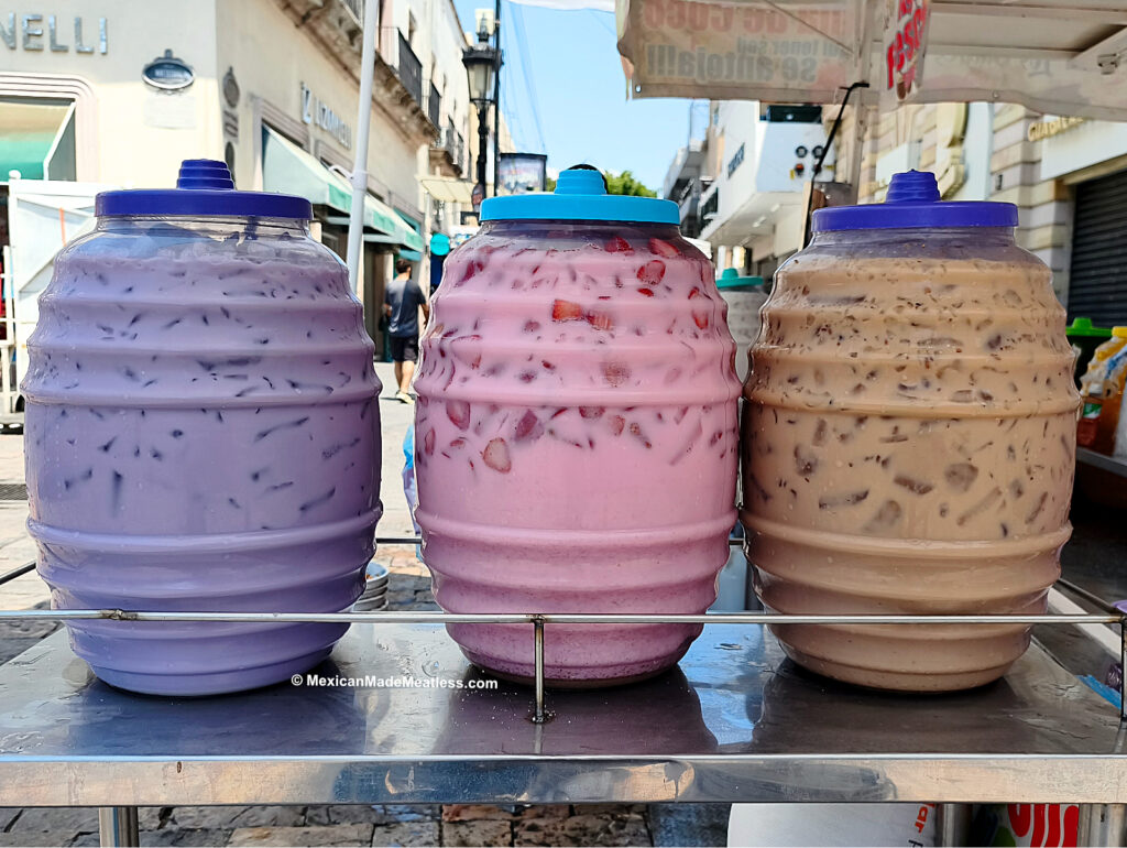 Three large plastic barrels filled with colorful agua fresca flavors—one purple, one pink, and one brown—are displayed on a street vendor’s stand in a bustling city setting.