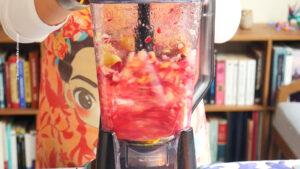 A close-up of a blender mixing colorful fruit pieces for guava agua fresca with beet, with a person in a vibrant, artistic apron standing behind it. Bookshelves are visible in the background.