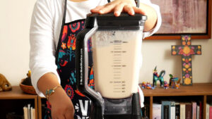 A person wearing a colorful apron blends a light-colored carlota de limon cream sauce in a large blender. Their hand is on the lid, and the background includes shelves with books, figurines, and a decorative cross.