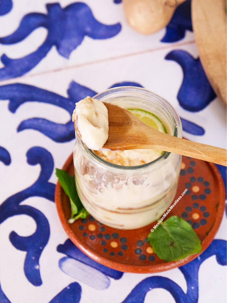 A wooden spoon with creamy white dairy free lime carlota dessert is held above a glass jar, which sits on a small terracotta plate with a basil leaf, on a decorative blue and white tiled surface.