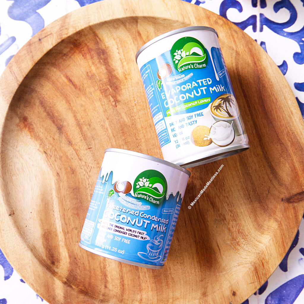 Two cans of Nature's Charm coconut milk products, one evaporated and one sweetened condensed, are placed on a round wooden tray with a blue-and-white patterned tile background.