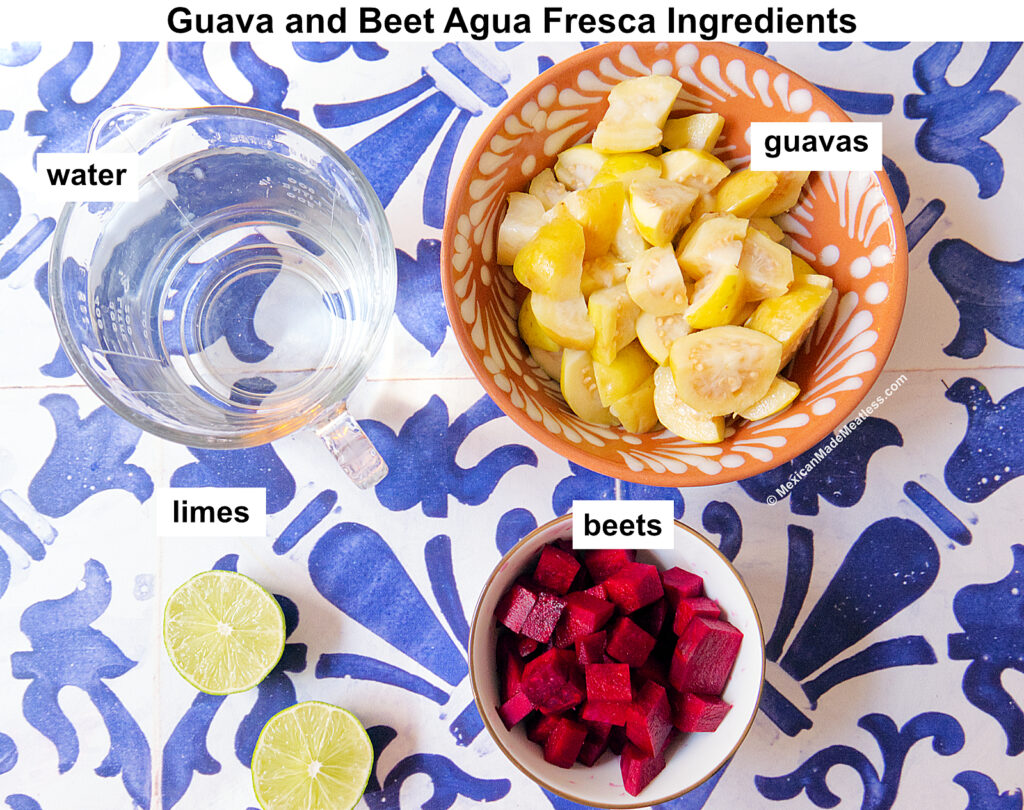 Overhead view of guava and beet agua fresca ingredients on a blue and white tile: a bowl of chopped guavas, a bowl of diced beets, a glass of water, and two cut limes. Ingredients are labeled.