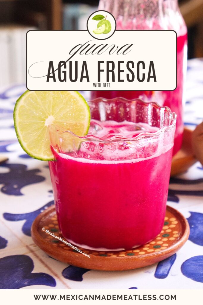 A glass of bright pink guava agua fresca with beet is garnished with a lime slice. The drink sits on a small clay coaster on a patterned table. A pitcher of the same drink is in the background.