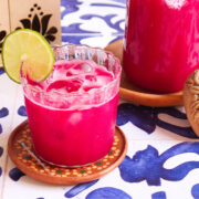 A glass of bright pink guava agua fresca with beet, filled with ice and a lime slice, sits on a patterned coaster next to a matching jug and decorative stone, all atop a blue and white tiled surface.