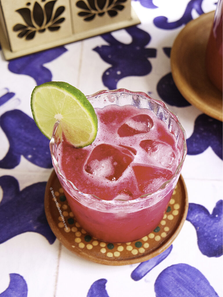 A vibrant pink guava agua fresca with beet, served over ice in a glass and garnished with a slice of lime. The glass rests on a decorative coaster atop a blue and white patterned tablecloth.