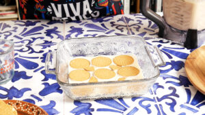 A glass baking dish with a layer of rectangular Maria cookies on a white creamy carlota de limon base, sitting on a blue and white tiled table. A blender with a beige mixture and someone in a "Viva Mexico" shirt are in the background.