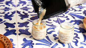 A blender pours a creamy carlota mixture into a small glass jar beside another filled jar, both on a blue and white patterned tile surface.