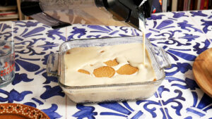 A blender pours a creamy dairy-free carlota de limon mixture over Maria cookies in a glass baking dish, resting on a blue and white patterned surface.