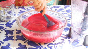A hand stirs bright pink guava agua fresca with beet in a glass bowl with a blue spatula on a blue and white tiled surface, next to a blender and a glass measuring cup.