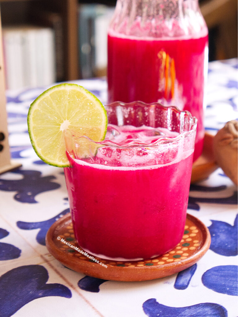 A glass of bright pink guava agua fresca with beet, garnished with a slice of lime, sits on a decorative coaster, with a pitcher of the same vibrant drink in the background on a blue and white patterned tablecloth.
