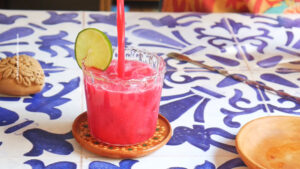 A bright pink guava agua fresca with beet is being poured into a glass with a slice of lime on the rim, set on a decorative coaster atop a blue and white patterned table. A clay ornament and wooden plate are visible nearby.