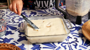 A hand spreads creamy white mixture over a glass baking dish filled with soaked bread, preparing a vegan carlota de limon next to a blender and a glass on a blue and white patterned table.