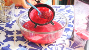 A bowl of bright red liquid is being strained through a black colander as seeds are separated with a blue spoon—perfect for straining guava seeds from agua fresca—on a blue-and-white patterned tile countertop.