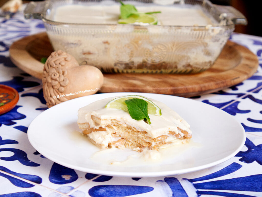 A slice of creamy vegan carlota de limon, topped with a lime wedge and mint, sits on a white plate; the rest of the dessert is in a glass dish in the background on a wooden board.