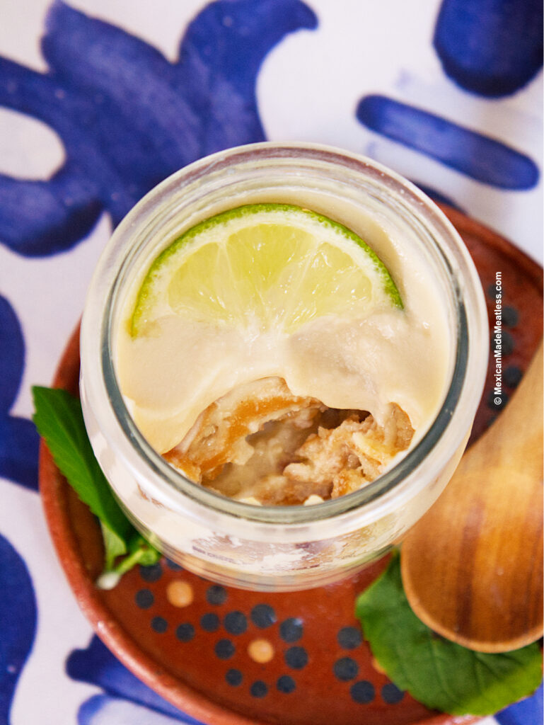 A small jar of vegan carlota de limon, creamy and partially eaten to reveal its layered texture, is topped with a lime slice. It sits on a brown dish with fresh green leaves and a wooden spoon nearby, on a blue and white patterned surface.
