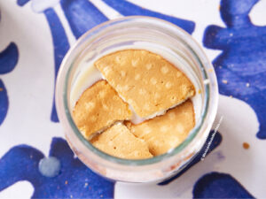 A glass jar with a layer of creamy vegan carlota de limon being layered in a jar, topped with several broken pieces of Maria cookies, placed on a blue and white patterned surface.