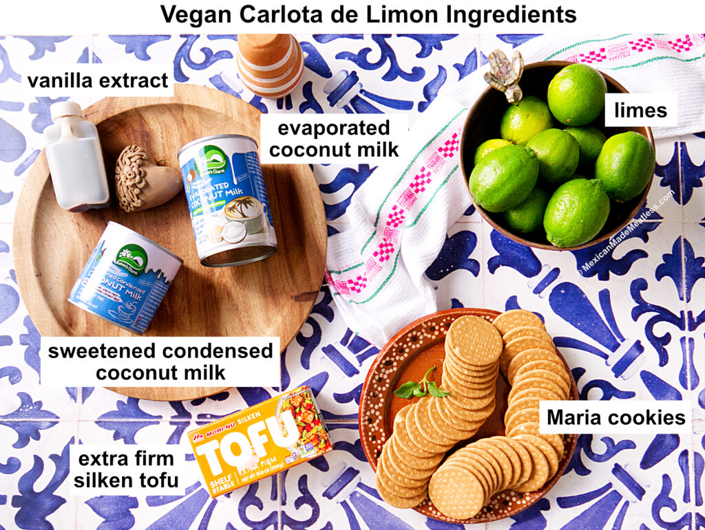Ingredients for Vegan Carlota de Limon on a blue and white tile background: vanilla extract, sweetened condensed coconut milk, evaporated coconut milk, extra firm silken tofu, limes, and Maria cookies.