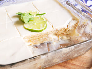 A glass baking dish with a vegan carlota de limon made with tofu, topped with a white creamy layer, garnished with two lime slices and a sprig of mint. One slice has been removed, revealing the dessert’s luscious layers inside.