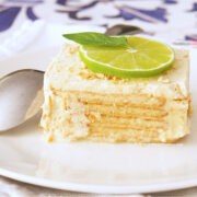 A square slice of creamy vegan carlota de limon, topped with a lime slice and mint leaves, sits on a white plate with a spoon beside it. The dessert features luscious layers and a light, crumbly topping.
