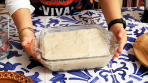 A person holds a glass baking dish filled with creamy carlota de limon ready to go in the refrigerator, covered with plastic wrap. The dish sits atop a blue and white patterned table, while the person sports a colorful apron.