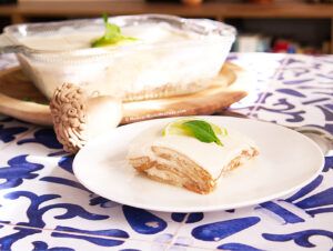 A slice of creamy dairy-free carlota de limon topped with lime and mint sits on a white plate, with the rest of the dessert in a glass dish in the background, all placed on a blue and white patterned surface.