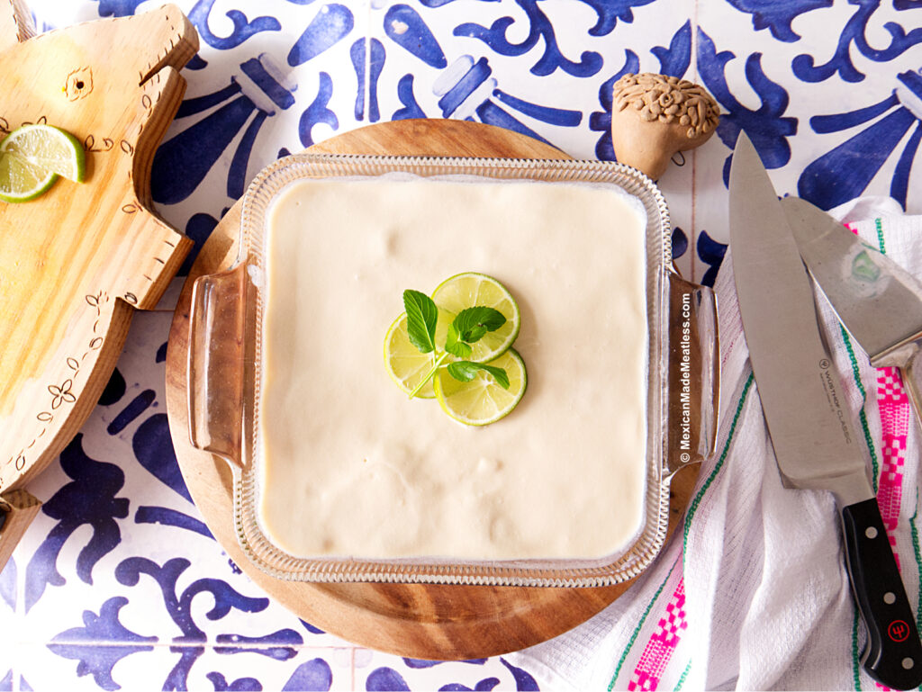 A square glass dish filled with creamy Carlota, Mexican no-bake dessert made with Maria cookies, garnished with lime slices and mint leaves, sits on a wooden board. Nearby are a large knife, an animal-shaped tray, and a white towel on a blue patterned surface.
