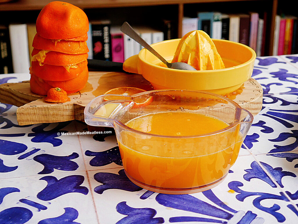 A bowl of fresh orange juice for naranjada sits on a blue and white tiled table, with a citrus juicer, spoon, and a stack of peeled oranges on a wooden board in the background. Books are visible on a shelf behind.