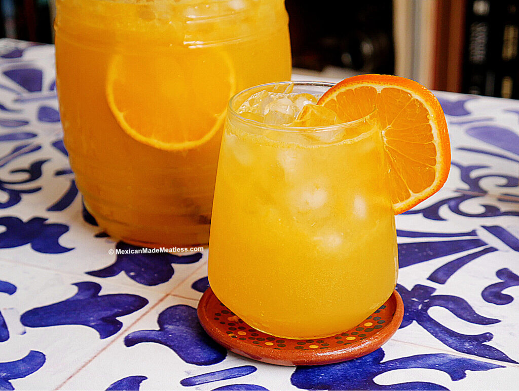 A glass of iced naranjada, a Mexican sparkling orange drink, garnished with an orange slice sits on a decorative coaster. A pitcher of the same vibrant drink is in the background on a blue and white patterned surface.