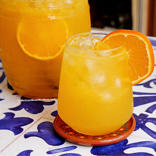 A glass of iced naranjada, a Mexican sparkling orange drink, garnished with an orange slice sits on a decorative coaster. A pitcher of the same vibrant drink is in the background on a blue and white patterned surface.