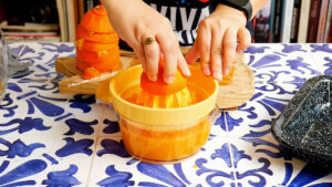 A person uses a manual orange juicer on a blue and white patterned table, pressing an orange half while stacked peels and a tray sit nearby, juicing oranges to make Mexican orangeade.