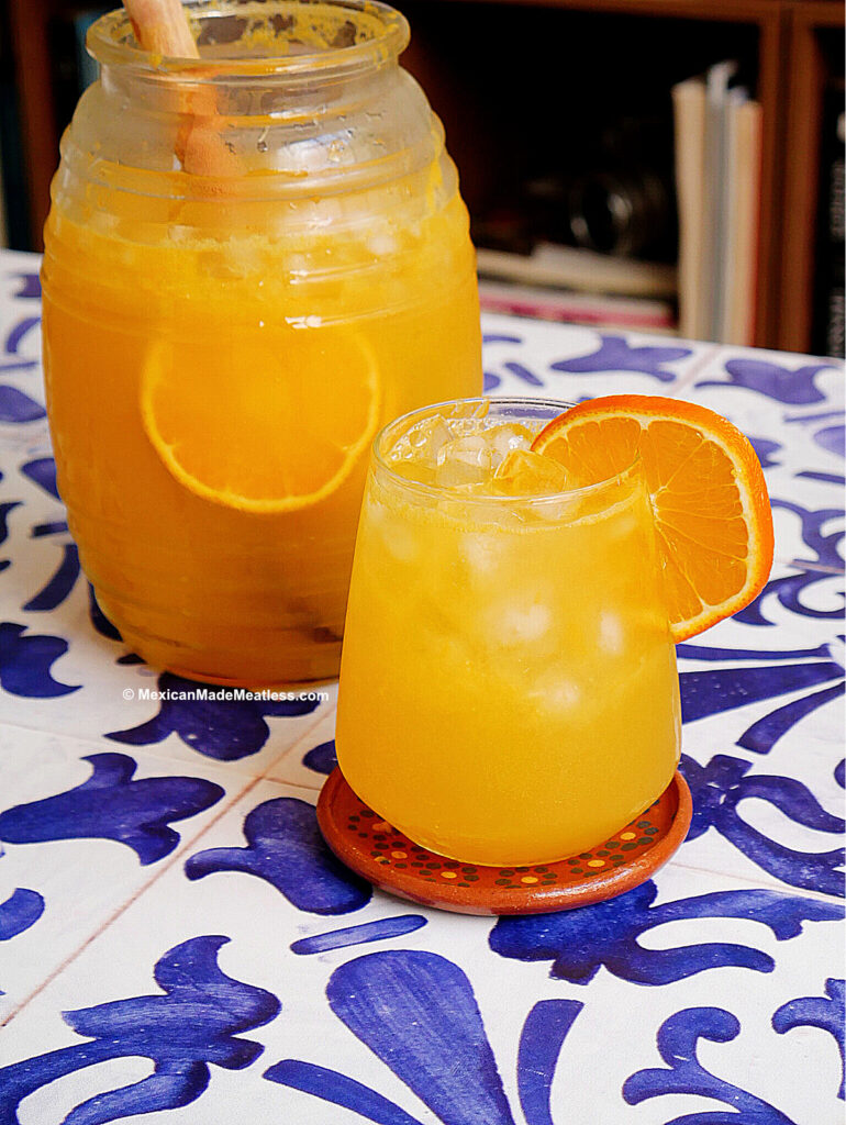 A glass and jug of naranjada, a sparkling orange agua fresca with ice and orange slices, sit on a blue-and-white patterned table; the glass is garnished with an orange slice.