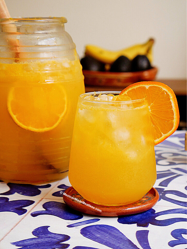 A glass of naranjada orange drink with ice and an orange slice garnish sits on a patterned blue and white table beside a large pitcher filled with the same drink; bananas and avocados are in the background.
