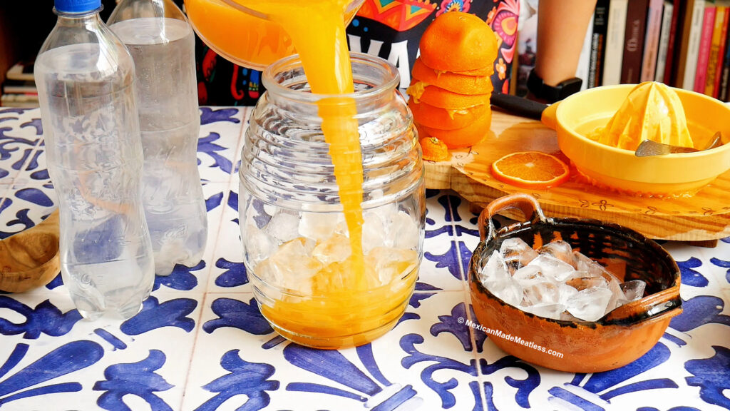 A person pours fresh orange juice, perfect for making naranjada Mexican sparkling orange drink, from a pitcher into a glass jar filled with ice on a blue and white tiled table surrounded by water bottles, a bowl of ice, oranges, and a citrus juicer.