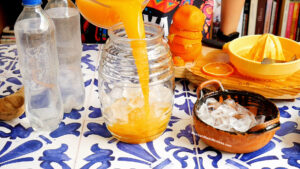 A person pours fresh orange juice, perfect for making naranjada Mexican sparkling orange drink, from a pitcher into a glass jar filled with ice on a blue and white tiled table surrounded by water bottles, a bowl of ice, oranges, and a citrus juicer.