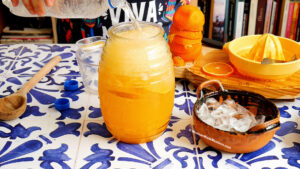A person pours water into a large glass jar filled with orange naranjada, surrounded by a wooden spoon, sliced oranges, a citrus juicer, a bowl of ice cubes, and bottle caps on a patterned blue and white table.