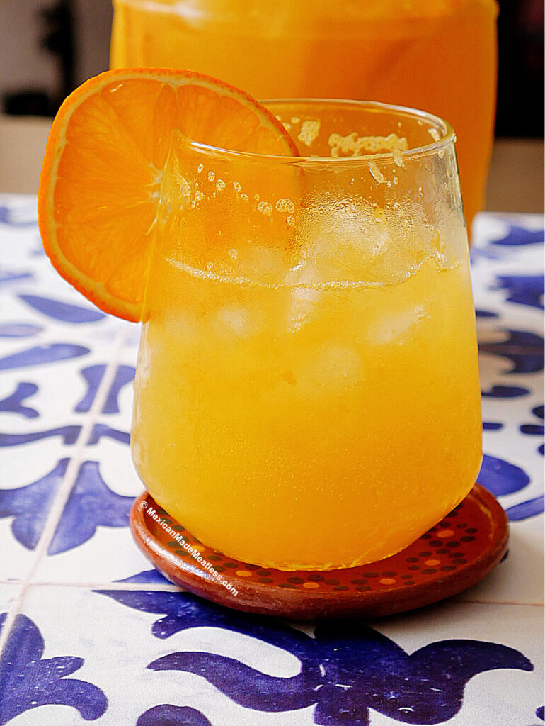 A glass of sparkling orange agua fresca without sugar with ice is garnished with an orange slice, sitting on a round coaster atop a blue and white patterned surface.