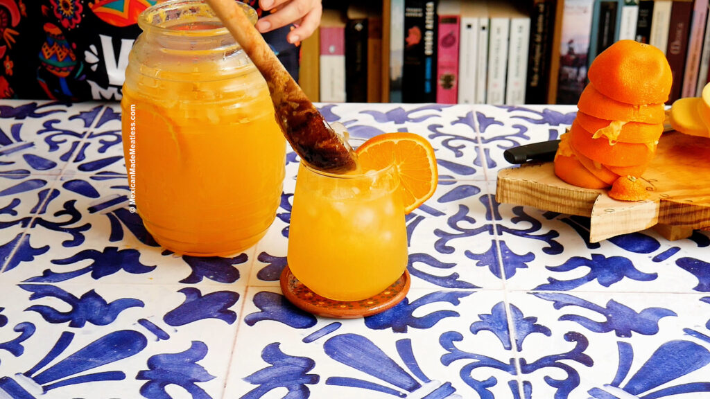 A glass of naranjada with ice and an orange slice sits on a blue-and-white tiled table next to a large pitcher of naranjada. A person stirs the jar, and peeled oranges are stacked on a wooden board nearby.