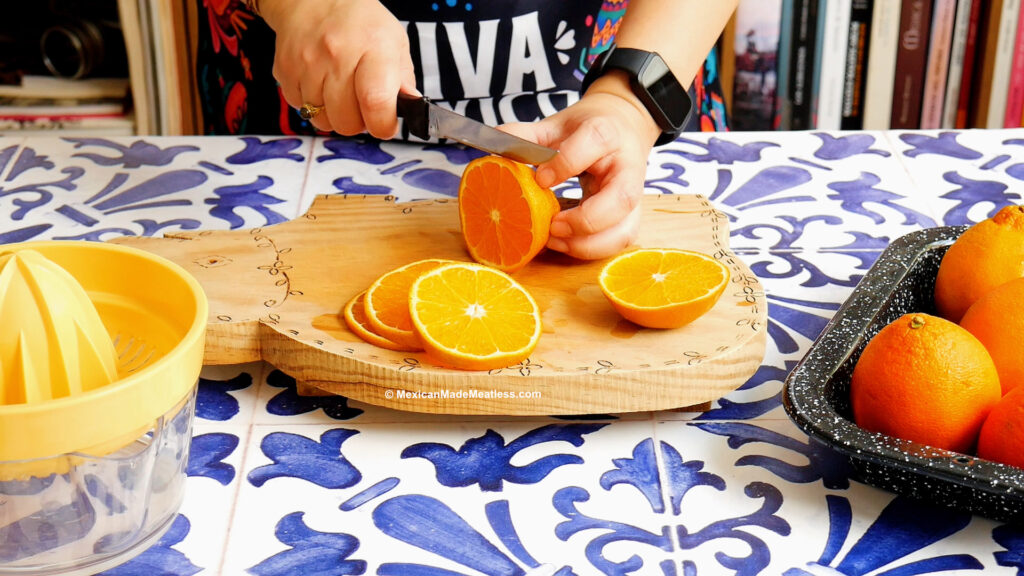 A person slices fresh oranges for naranjada on a wooden cutting board atop a blue and white tiled counter, with whole oranges in a tray and a citrus juicer nearby.