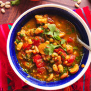 A colorful bowl of vegan frijoles charros, made with beans, vegan hot dogs, and vegetables and garnished with fresh cilantro, sits on a red woven placemat, with a spoon inside and beans scattered on the wooden table.