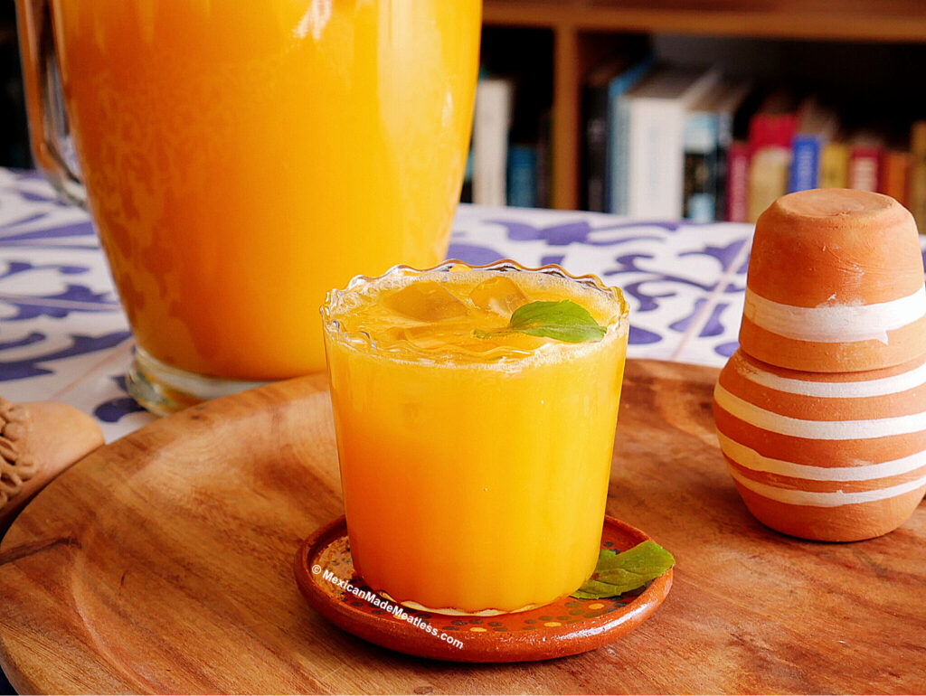 A glass of mango agua fresca with ice and mint leaves sits on a wooden tray, with a pitcher of agua de mango and a striped clay cup in the background.