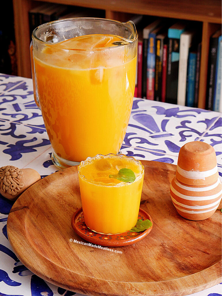 A glass and pitcher of refreshing mango agua fresca with ice sit on a wooden tray, garnished with mint. The background features a patterned blue-and-white tablecloth, a clay cup, and a bookshelf.