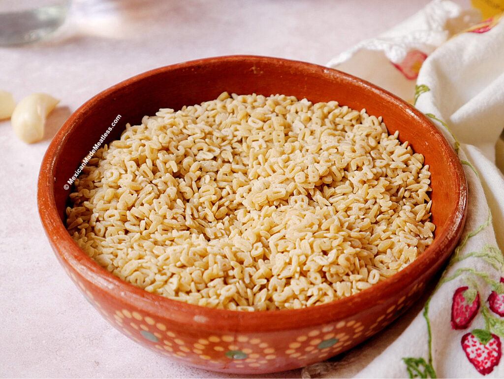 A round, barro bowl filled with cooked alphabet pasta to make Mexican sopa de letras, placed on a light surface next to a white and floral-patterned napkin.