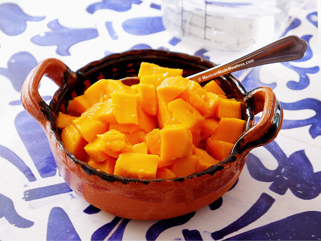 A rustic brown clay bowl filled with bright yellow Ataulfo mango cubes sits on a blue and white patterned surface, with a spoon resting inside the bowl. A glass measuring cup is in the background.