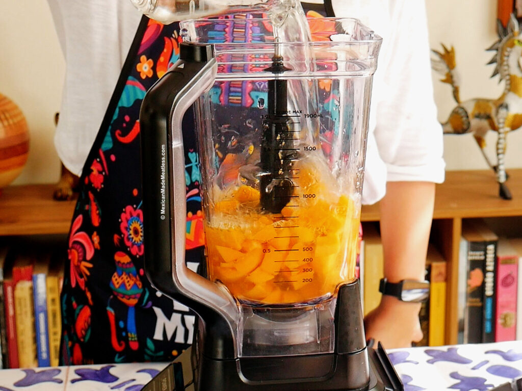 Nancy Lopez is wearing a colorful apron and pouring water into a blender filled with chopped mangoes. The blender sits on a table with a blue floral tablecloth, and bookshelves with decorative items are in the background.
