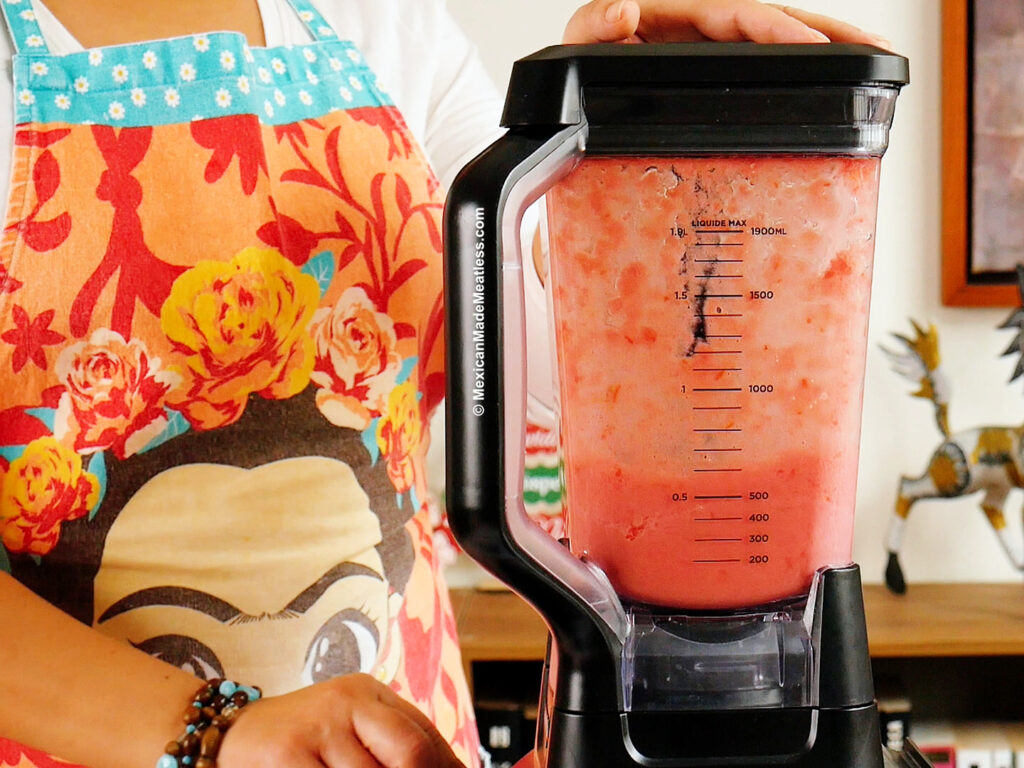 A person wearing a colorful floral apron blends a tomato mixture in a large blender, to prepare tomato sauce for Mexican alphabet soup. Their hand rests on the lid, and measurement markings are visible amid the vibrant kitchen setting.