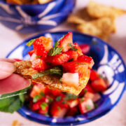 A hand holds a cinnamon sugar tortilla chip topped with strawberry mint salsa. In the background, a bowl of the same salsa and chips are visible, along with a whole strawberry.